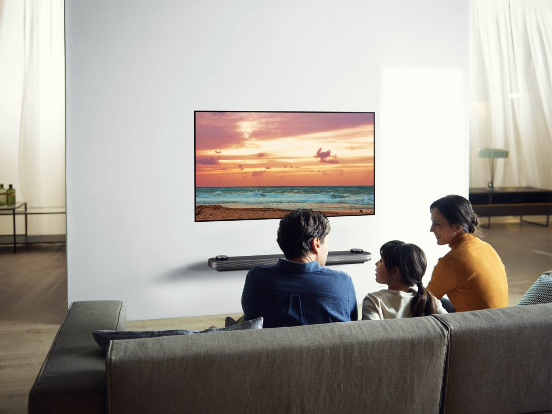 Another view of a family sitting in their living room looking at the LG SIGNATURE OLED TV W (model 7) which is mounted seamlessly on the wall