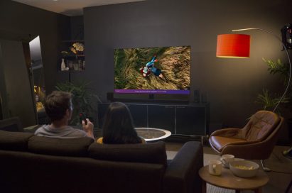 A man and woman seated on a couch in their living room at night watching a couple laying in long grass on their LG AI OLED TV