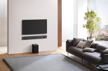 An upper view of the LG Soundbar model SL9YG installed on the wall below an LG TV, with the LG Wireless Rear Speaker Kit model SPK 8 on the floor below